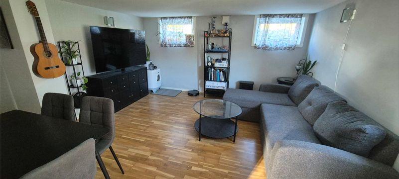 Stue med grå sofa, glassbord, TV, gitar på veggen og bokhylle i et lyst rom med parkettgulv.