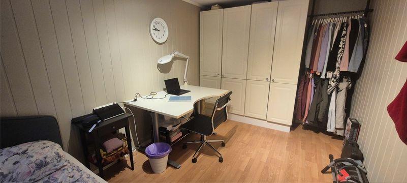 Et rom med skrivebord, stol, lampe, og skap, samt en klesstang med klær og en seng i bakgrunnen.