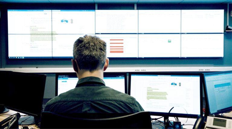En person sitter foran flere dataskjermer i et kontrollrom, med fokus på informasjon på skjermene.