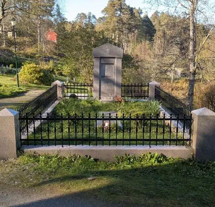 Gravsted med steinmonument og jerngjerde, omkranset av grønt gress og blomster i en park.