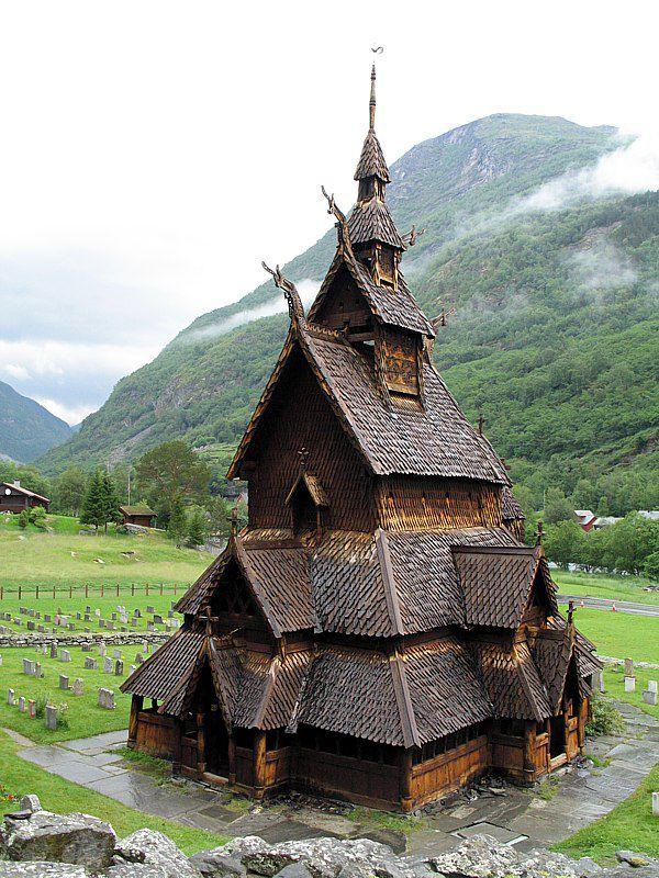 Stavkirke i norsk stil med spisse tårn, omgitt av fjell og en gravplass i bakgrunnen.