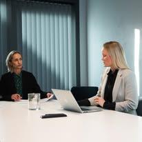 To kvinner i et møterom, en med laptop og dokumenter, den andre lytter oppmerksomt.