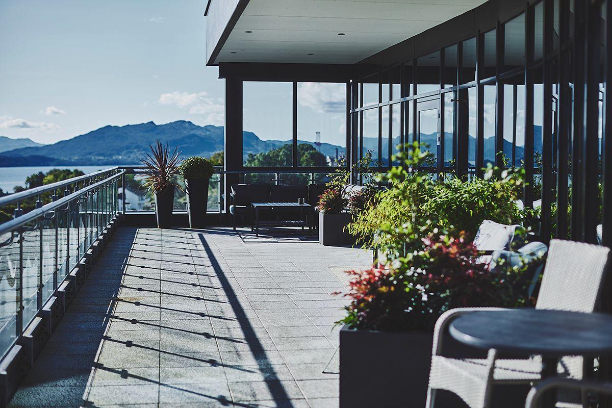 En moderne terrasse med blomster og utsikt over fjellene og vannet. Sollys skaper skygger på gulvet.
