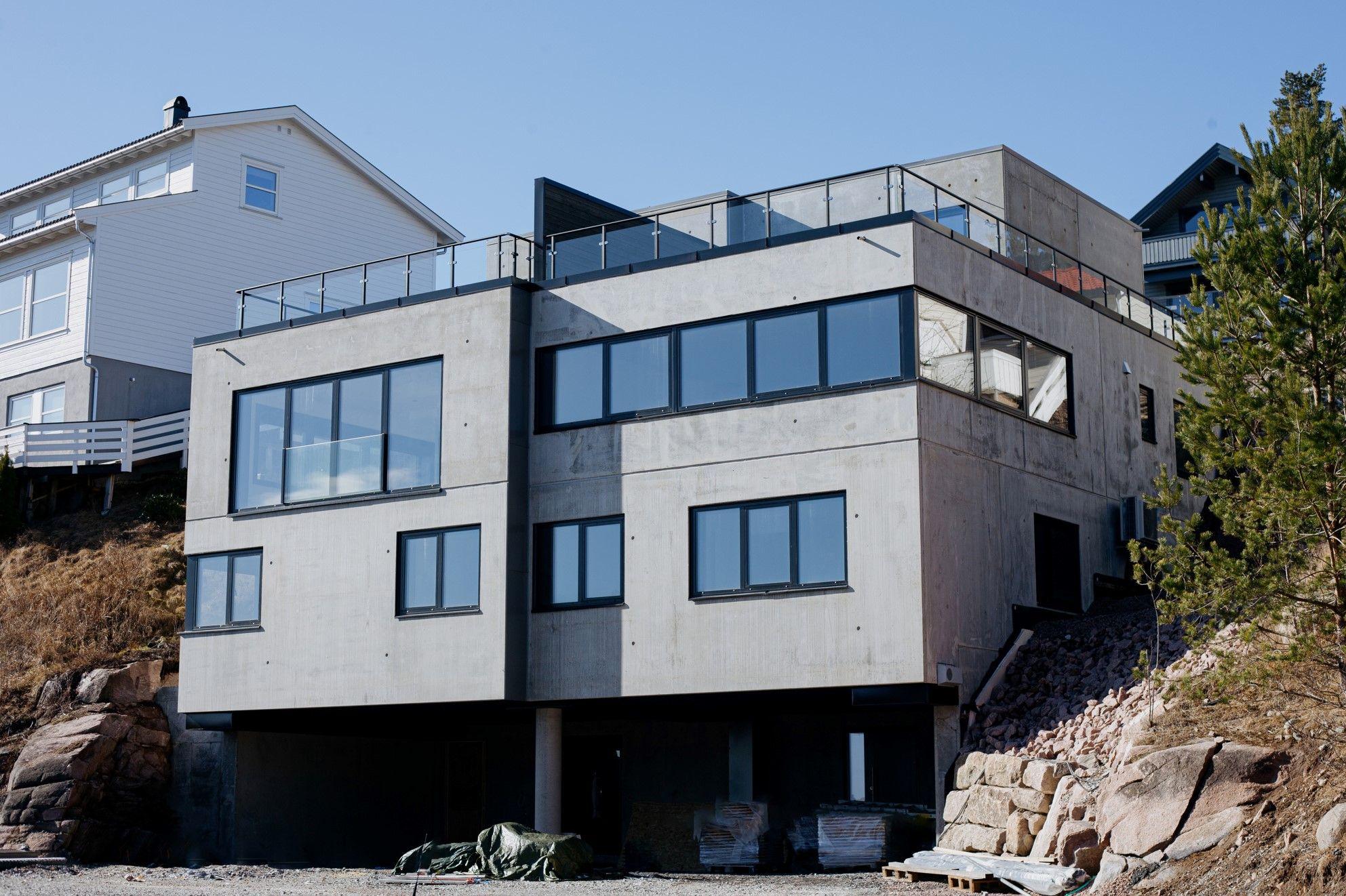 Moderne betonghus med store vinduer og takterrasse, beliggende på en skråning.