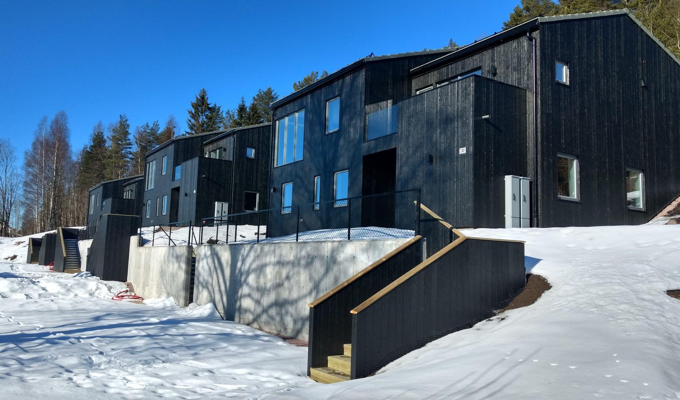 Moderne, svarte hus med store vinduer, omgitt av snø og trær i en solfylt vinterlandskap.