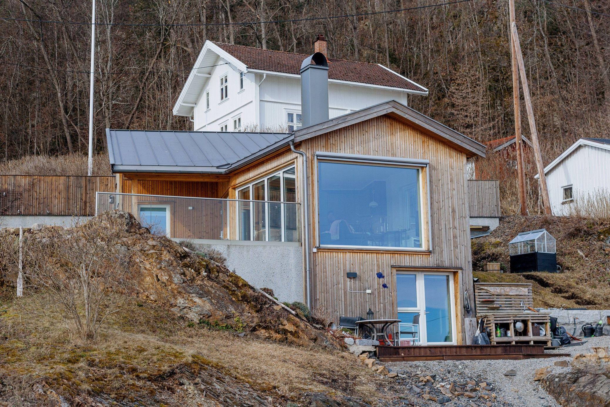 Moderne hytte med trevegg og store vinduer, beliggende på en skråning med utsikt over landskapet.