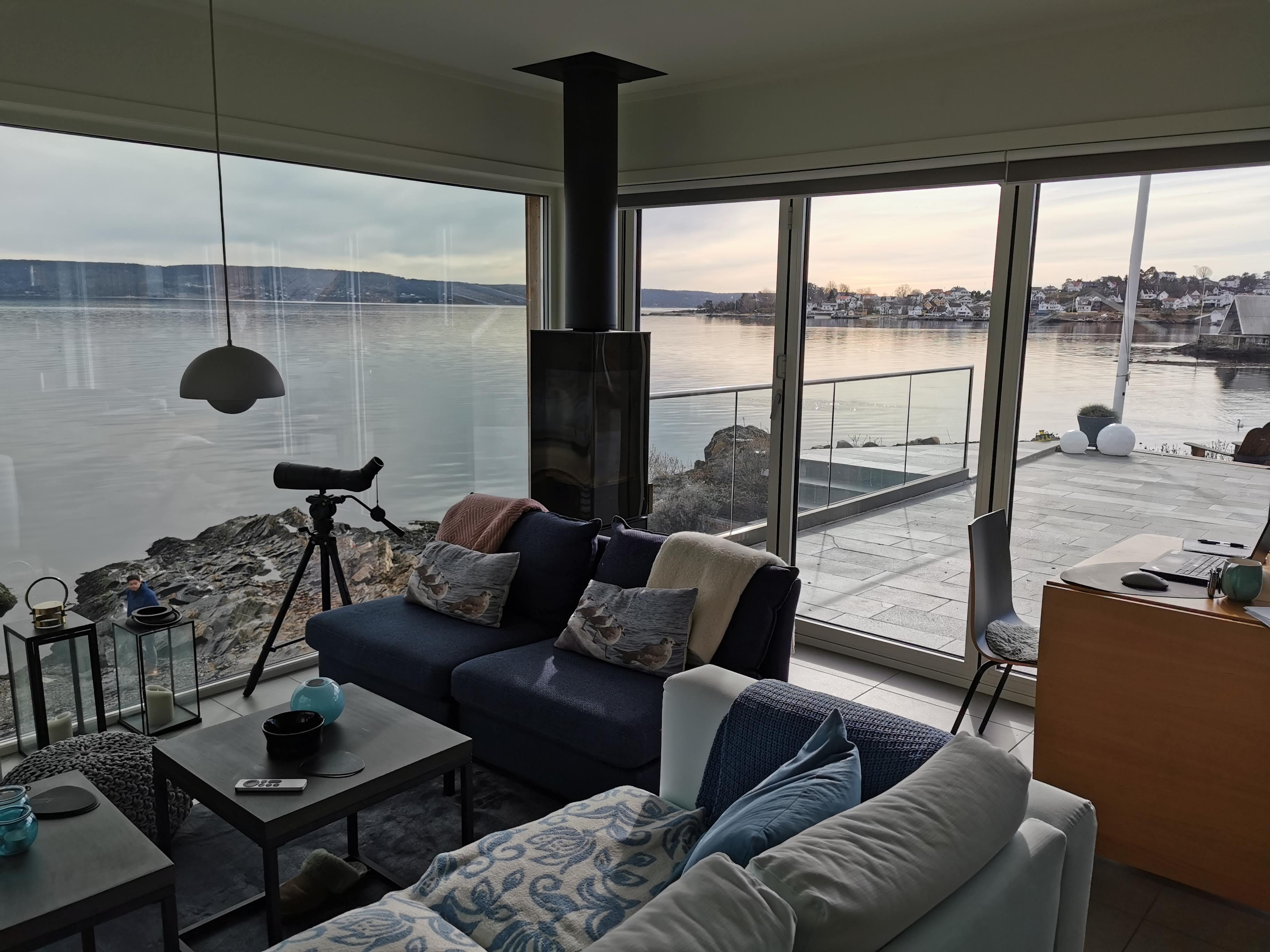 Moderne stue med utsikt over sjøen, store vinduer, sofaer, og et teleskop i et lyst rom.