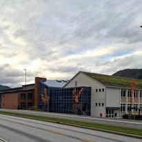 Moderne skolebygning med grønt tak og store vinduer, omgitt av fjell og en grå himmel.