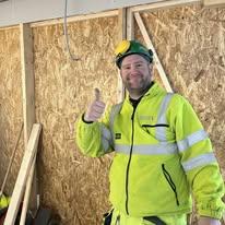 En smilende mann i gul arbeidsjakke og hjelm gir tommel opp på en byggeplass med treplanker i bakgrunnen.