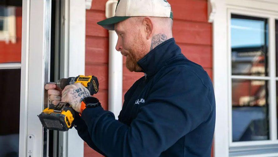 En mann med tatoveringer bruker en drill på en rød husvegg.