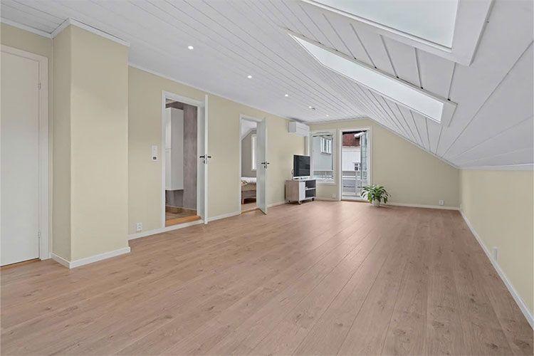 Lyst og romslig loftstue med tregulv, vinduer og utgang til balkong.