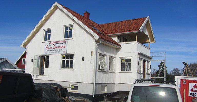 Hvit enebolig med rød takstein og balkong, under oppussing, med byggeutstyr i forgrunnen.