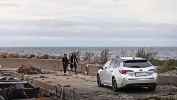 To kvinner går langs en brygge ved havet, med en hvit bil parkert i forgrunnen.