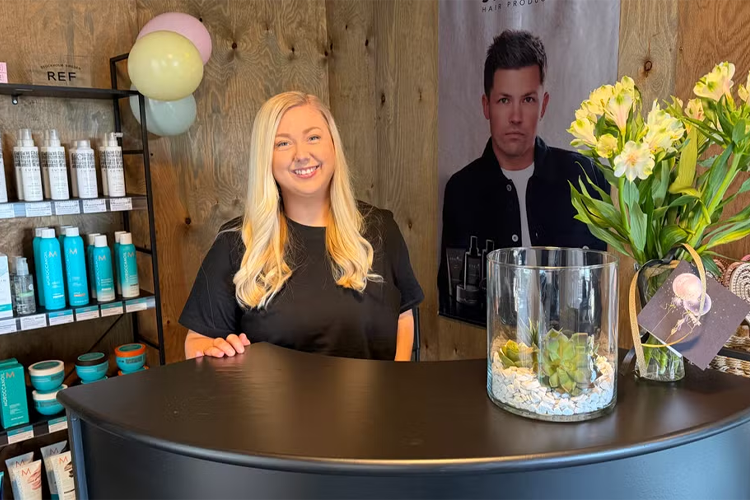 En smilende kvinne står bak en resepsjon med hårprodukter og blomsterdekorasjon.