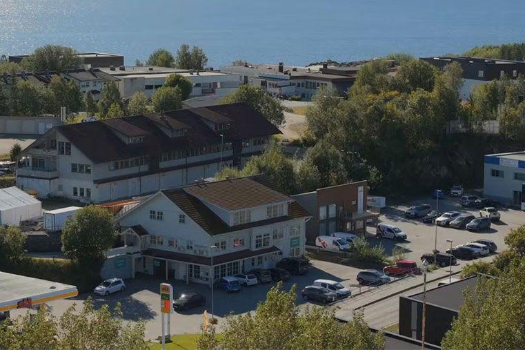 Utsikt over en by med bygninger, parkerte biler og havet i bakgrunnen. Grøntområder omgir området.