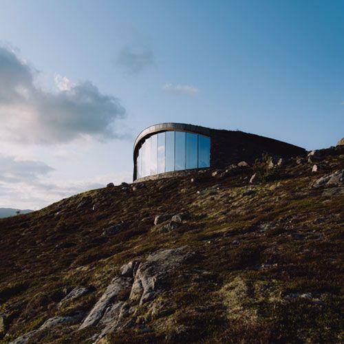 Moderne bygning med store vinduer på en fjellside, omgitt av steiner og lyng under en blå himmel.
