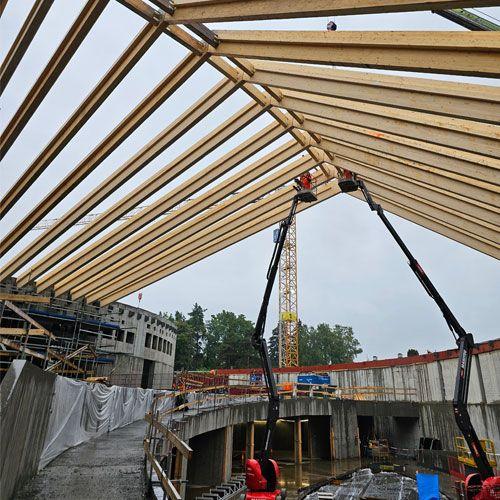 Byggeplass med takstoler under konstruksjon, kran og arbeidere i regnvær.