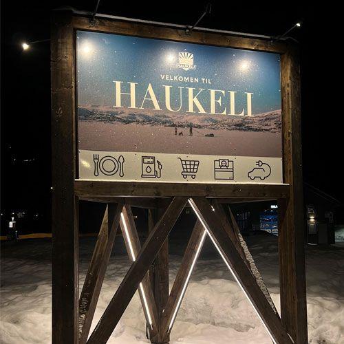 Velkomstskilt til Haukelifjell med ikoner for mat, shopping og bil, omgitt av snø om natten.