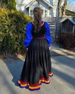 Kvinne iført tradisjonell bunad med blå ermer og svart skjørt, står med ryggen til foran et hus.