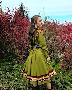 Kvinne i tradisjonell samisk drakt står foran blomstrende busker i naturen.
