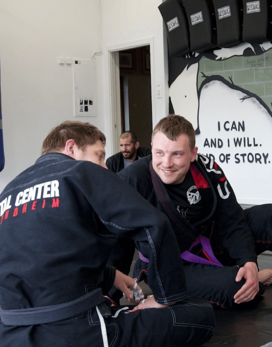 To personer i jiu-jitsu drakt smiler og snakker sammen på treningsmatten i en dojo.