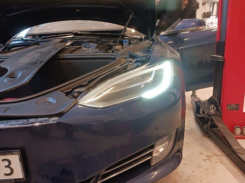 En blå Tesla med åpen motorlokk og lysene på, står i et verksted.