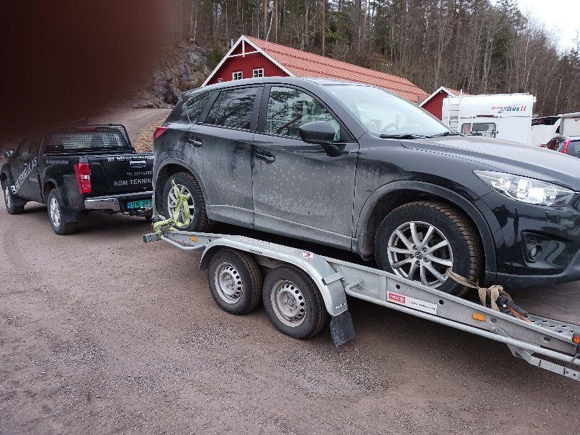 En svart SUV på en tilhenger med en grå pickup i bakgrunnen, parkert på en grusvei.