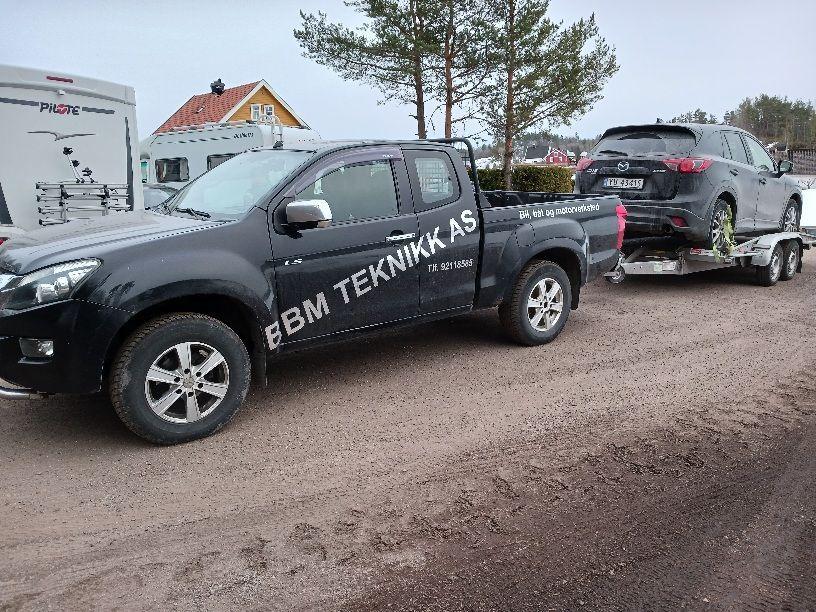 En svart pickup med påhenger som frakter en bil, med teksten "EBM TEKNIKK AS" på siden.