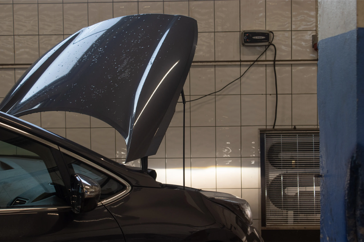 En bil med åpen motorlokk står i en bilvask, med fliser og ventilasjonsanlegg i bakgrunnen.