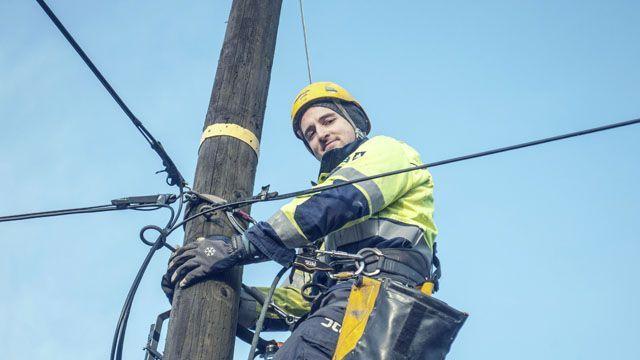 En elektriker klatrer opp en stolpe mens han arbeider med ledninger under en klar blå himmel.