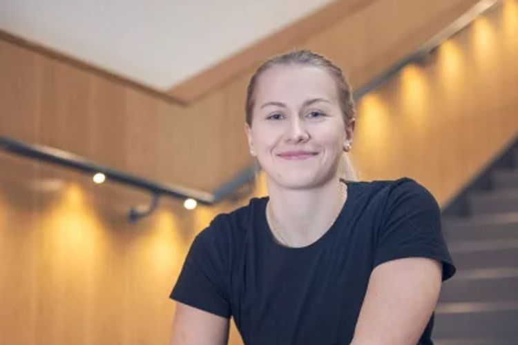 En smilende kvinne med kort hår sitter på en trapp med trevegger og dempet belysning i bakgrunnen.