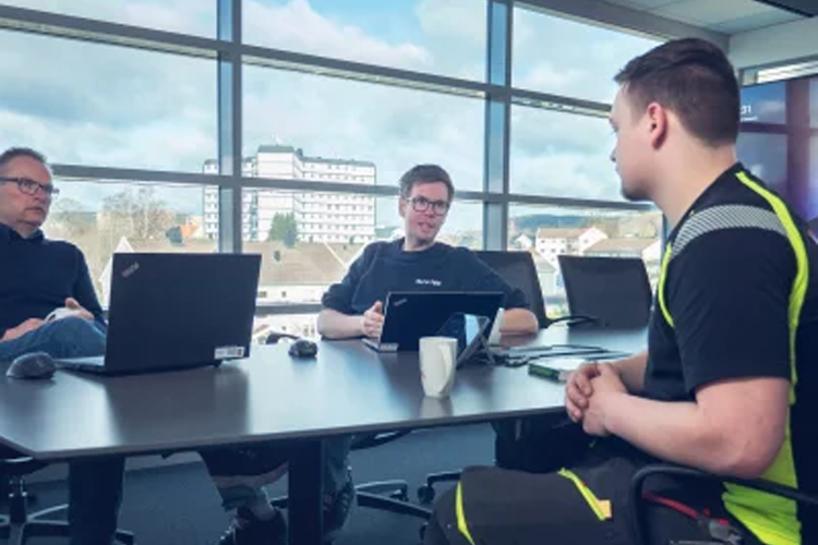 Tre menn sitter rundt et bord med bærbare datamaskiner, diskuterer i et moderne kontor med vinduer som viser utsikt.