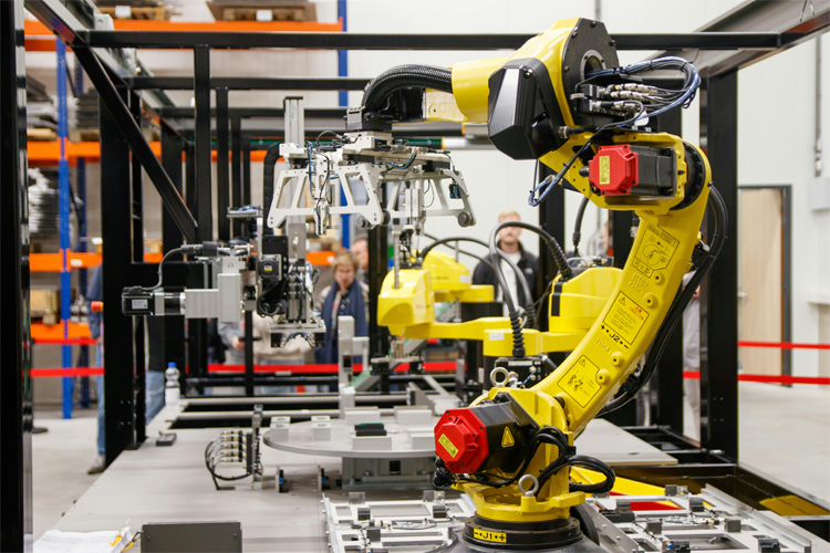 Industriell robotarm i en fabrikk med mennesker i bakgrunnen som observerer produksjonsprosessen.