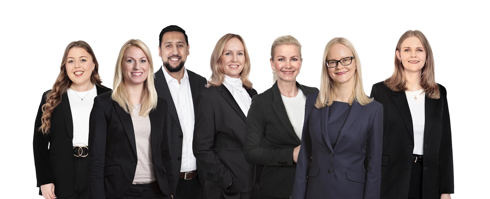 En gruppe på syv profesjonelle personer i formelle klær, smilende og poserende sammen foran en hvit bakgrunn.