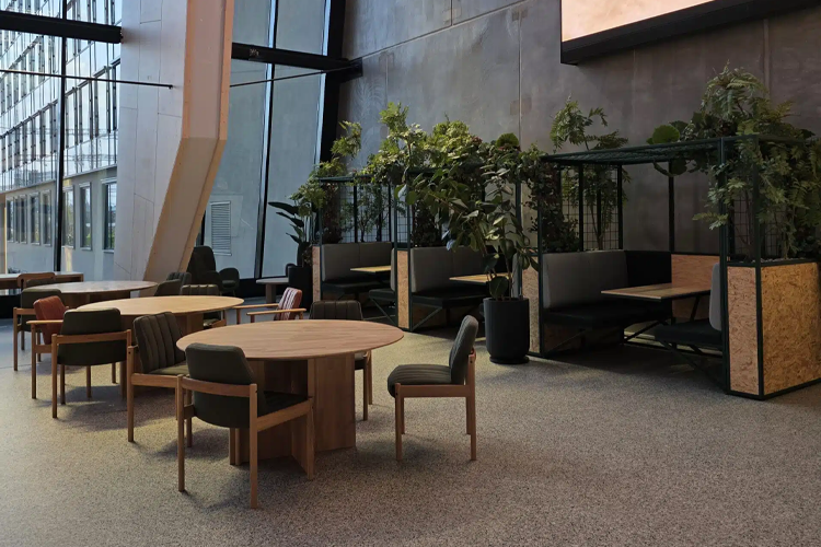 Moderne kontorlandskap med runde bord, stoler og grønne planter i et lyst rom med store vinduer.