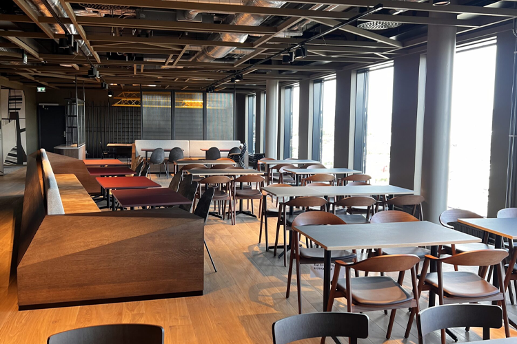 Moderne restaurantinnendørs med trebord og stoler, store vinduer og en lys atmosfære.