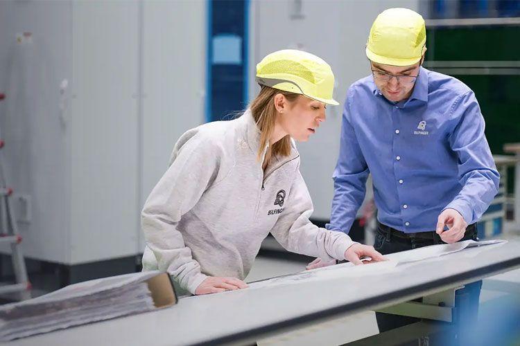To personer i sikkerhetshjelmer ser på dokumenter i en fabrikk. En kvinne og en mann samarbeider om et prosjekt.