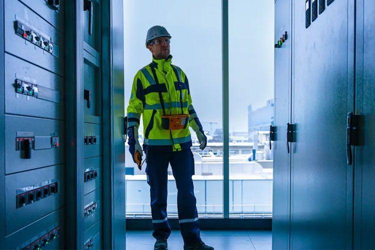 En arbeidstaker i sikkerhetsutstyr står mellom kontrollskap i et moderne bygg.