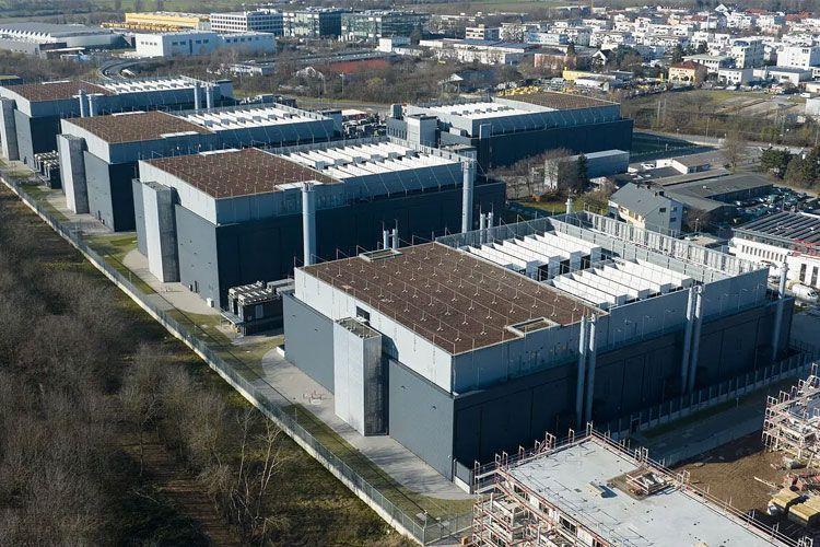 Moderne industribygg med flate tak og solcellepaneler, omgitt av grønt område og bylandskap i bakgrunnen.