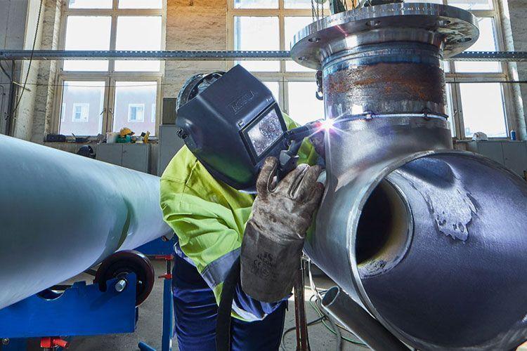 En sveiser som arbeider med rør i en industriell setting, iført verneutstyr og sveisehjelm.