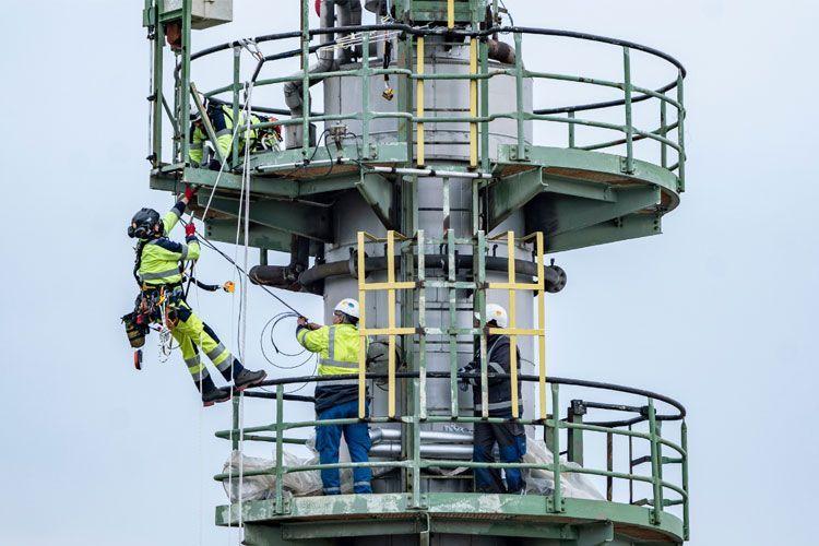 Arbeidere i sikkerhetsutstyr klatrer opp en høy mast med tau og utstyr for inspeksjon og vedlikehold.