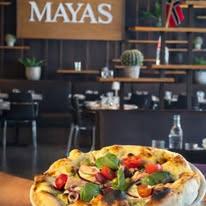 En person holder en pizza med grønnsaker og urter, med restauranten Mayas i bakgrunnen.