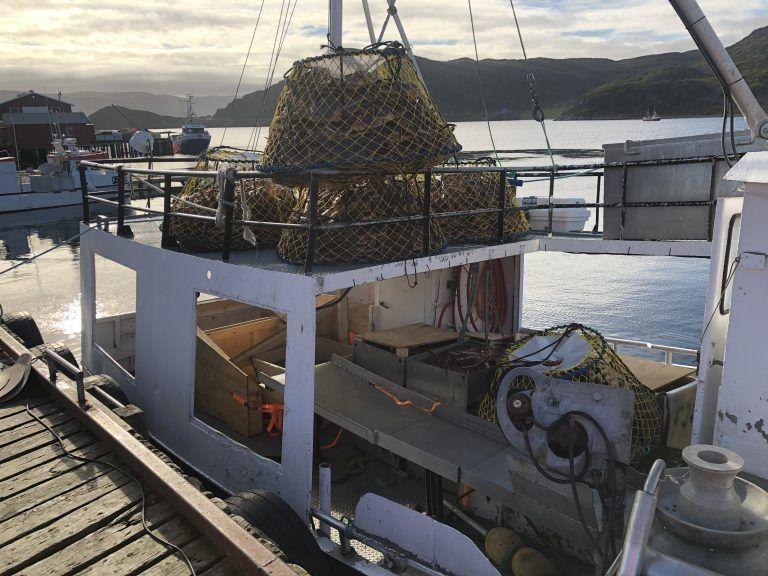 Fiskebåt med fiskeutstyr og garn på dekk, liggende ved kai i en rolig havn.