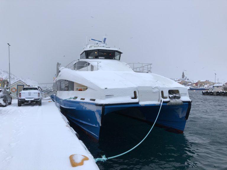 En stor katamaranbåt dekket av snø ved havnen, med en bil parkert i bakgrunnen.