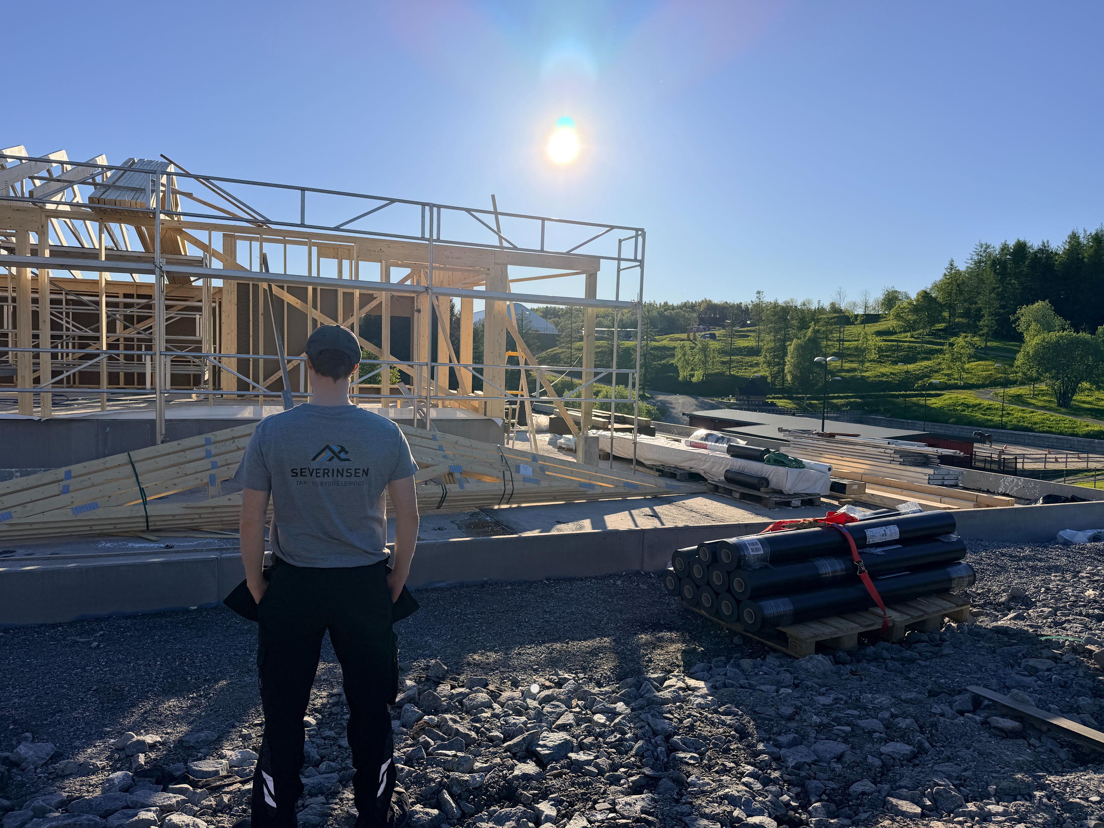 Bygningsarbeider ser på byggeplassen med soloppgang i bakgrunnen og materialer stablet på bakken.