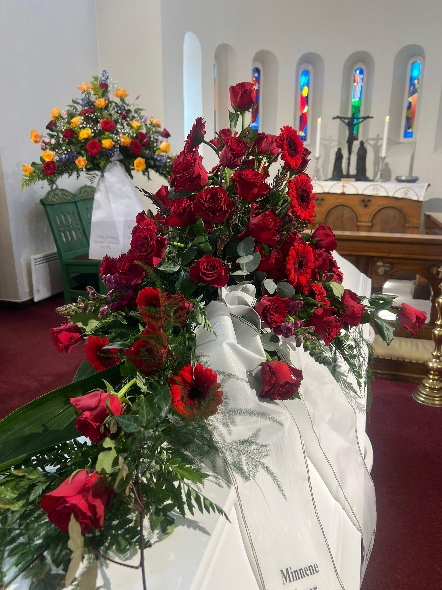 En hvit kiste dekorert med røde roser og blomster i en kirke med fargerike vinduer.
