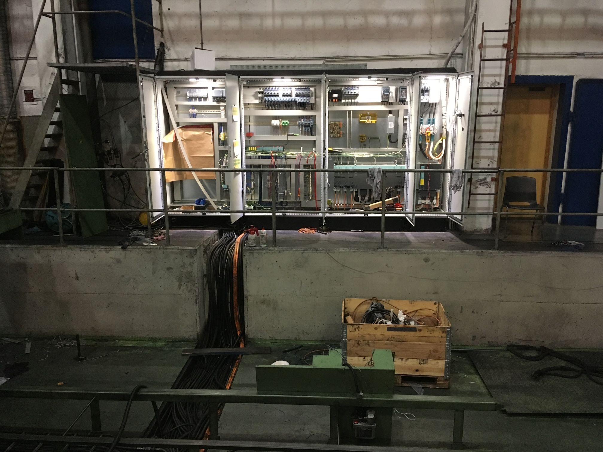 En industriell innendørs scene med elektriske skap, kabler og en trepakke på gulvet.