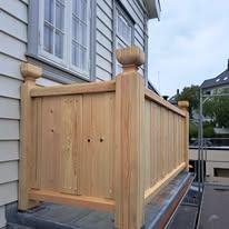 En treverksrekkverk med runde stolper, plassert på en bygning med grå tak og vinduer i bakgrunnen.