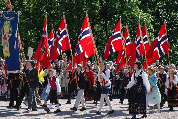 Barn og voksne i tradisjonelle klær går i 17. mai-tog med norske flagg og feststemning.