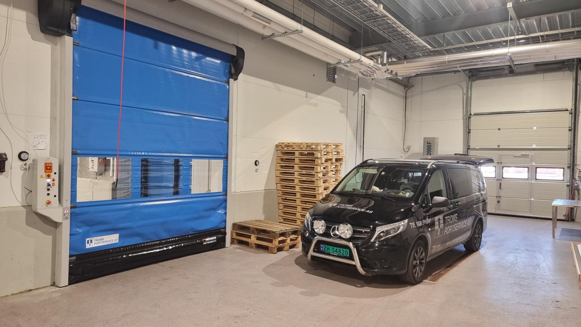 En svart varebil står ved en blå industridør i et lager med paller i bakgrunnen.